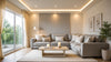 A cozy living room with a large gray sofa, white cushions, and a wooden coffee table with lit candles. Soft lighting, earth tones, and a large window creating a serene atmosphere.