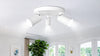 Ceiling-mounted white spotlight fixture with three adjustable lights, illuminating a bright room. Nearby are a window, leafy plant, and framed artwork.