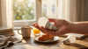 A hand holds an LED bulb over a wooden table with a mug, croissant, and juice. Another bulb lies nearby. Soft morning light streams through a window.