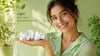 A smiling woman in a green blouse holds LED bulbs in her palm against a lush, plant-filled background. Text reads "Light that cares, for a better tomorrow."