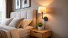 A cozy bedroom with a beige upholstered headboard, neatly arranged pillows, and a wooden nightstand with a small plant. A wall lamp emits warm light, creating a calming ambiance.